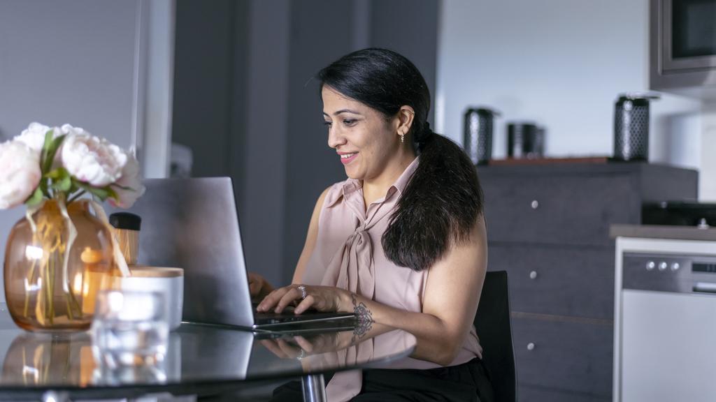 A beautiful woman of Indian descent smiles while using a laptop computer to shop online and do online banking. She is sitting at a table in her modern home.