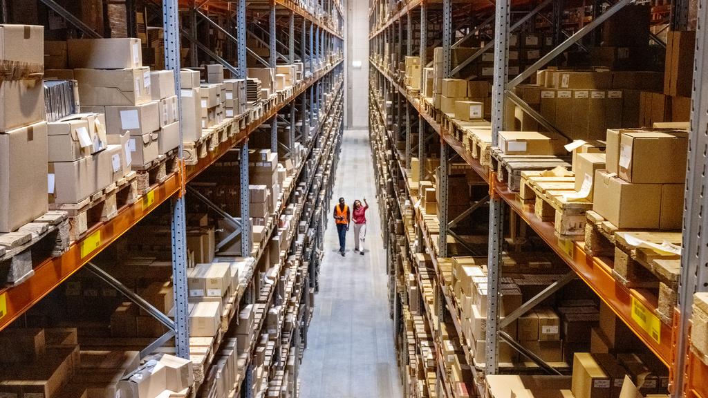 High angle view of a warehouse manager walking with foremen checking stock on racks. Businesswoman discussing stock with a male worker while walking by racks in warehouse.